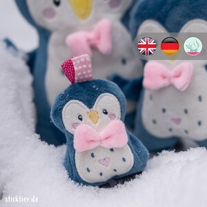 Könnte beinhalten: Ein blauer Pinguin-Plüschtier mit einer rosa Schleife und weißen Punkten. Der Pinguin sitzt im Schnee.