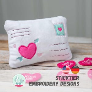 Peut inclure: Un coussin en peluche blanc en forme de lettre, brodé d'un cœur rose percé d'une flèche turquoise. Des détails supplémentaires de cœurs turquoise et roses et un motif de timbre-poste sont visibles. Le coussin fait partie de la collection Sticktier Embroidery Designs.