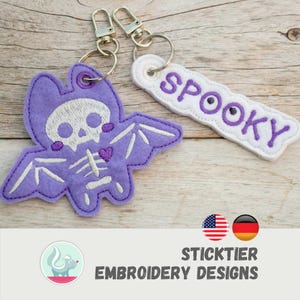 May include: Two Halloween keyrings. One is a purple bat with a skeleton design, and the other is a white tag with the word "SPOOKY" in purple. Both have silver key rings. The keyrings are on a wooden surface.