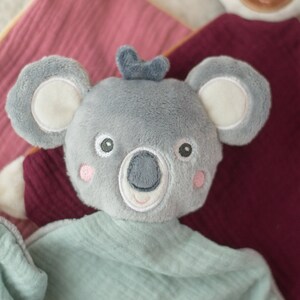 May include: A gray koala plush toy with a light blue shirt and pink cheeks. The koala has large ears with white inner ears and a small blue bow on its head. The toy is on a pink and burgundy fabric.