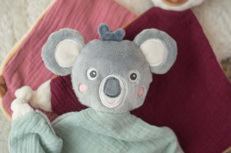 May include: A gray koala plush toy with a light blue shirt and pink cheeks. The koala has large ears with white inner ears and a small blue bow on its head. The toy is on a pink and burgundy fabric.