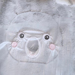 May include: A close-up of a baby blanket featuring a koala face design. The koala's face is embroidered with gray, white, and pink thread. The blanket has a soft, plush texture, perfect for a baby.