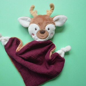 May include: A deer-shaped baby comfort blanket. The deer has a brown head with white ears and a burgundy blanket. The deer's face has embroidered details, including eyes, a nose, and rosy cheeks. The blanket is made of a soft, textured fabric.