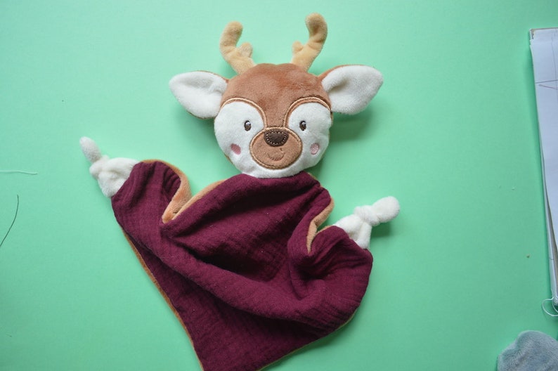 May include: A deer-shaped baby comfort blanket. The deer has a brown head with white ears and a burgundy blanket. The deer's face has embroidered details, including eyes, a nose, and rosy cheeks. The blanket is made of a soft, textured fabric.