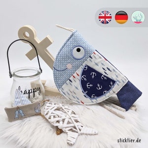 Puede incluir: Un pez de tela azul y blanco con un patrón de ancla y peces. El pez tiene un ojo grande y una boca rosa. El pez está sobre un fondo blanco con un ancla de madera y un frasco de vidrio con la palabra "happy" en él.