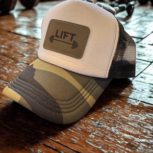 May include: A white and black mesh-backed trucker hat with a camouflage brim. The hat features a brown leather patch with the word "LIFT" and a barbell graphic. The hat is likely designed for gym or fitness enthusiasts.