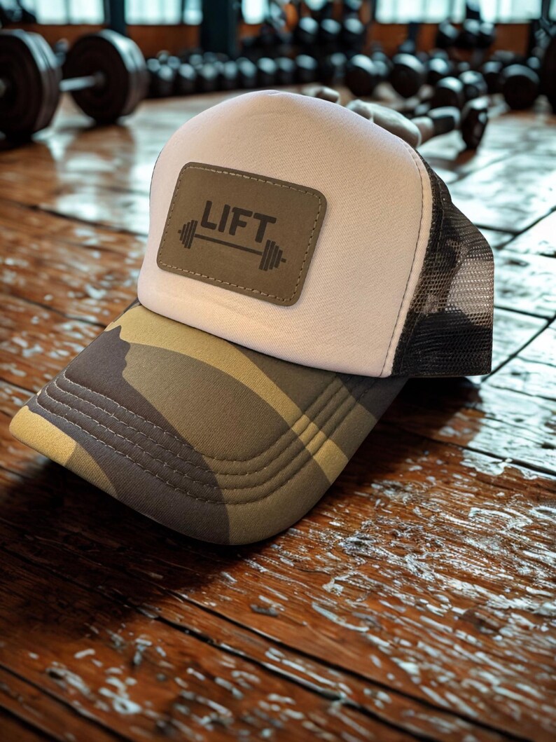 May include: A white and black mesh-backed trucker hat with a camouflage brim. The hat features a brown leather patch with the word "LIFT" and a barbell graphic. The hat is likely designed for gym or fitness enthusiasts.