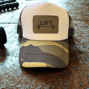 May include: A trucker hat with a white front panel, a camouflage brim, and a mesh back. The hat features a rectangular patch with the word "LIFT" and a barbell graphic. The hat is designed for fitness enthusiasts.
