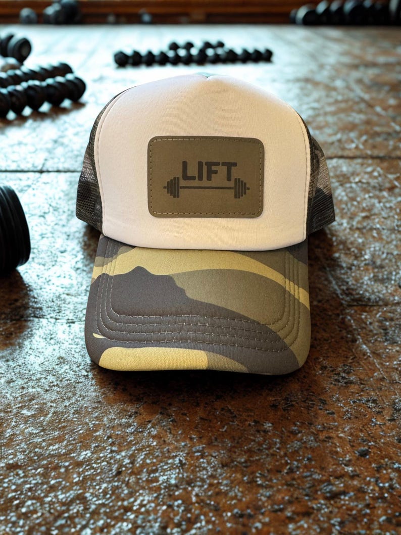 May include: A trucker hat with a white front panel, a camouflage brim, and a mesh back. The hat features a rectangular patch with the word "LIFT" and a barbell graphic. The hat is designed for fitness enthusiasts.