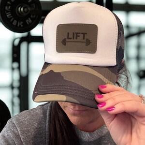 May include: A white and black mesh trucker hat with a camouflage brim. The hat features a rectangular patch with the word "LIFT" above a barbell graphic. The hat is being held up by a person with pink fingernails.