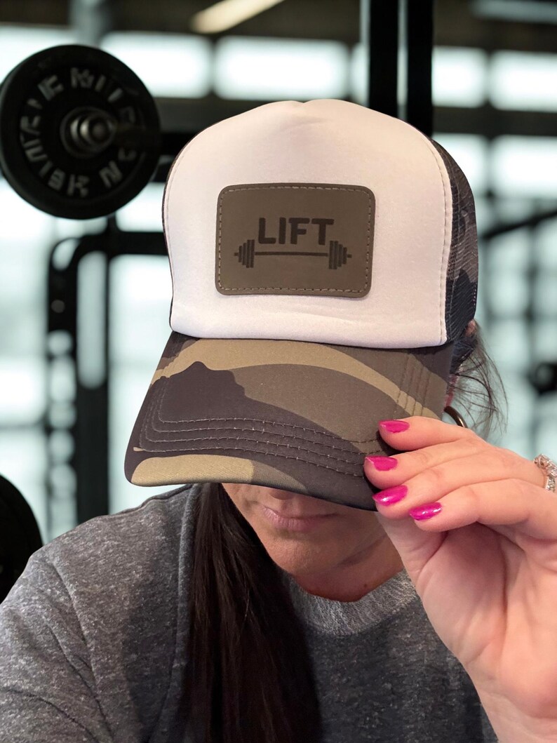 May include: A white and black mesh trucker hat with a camouflage brim. The hat features a rectangular patch with the word "LIFT" above a barbell graphic. The hat is being held up by a person with pink fingernails.