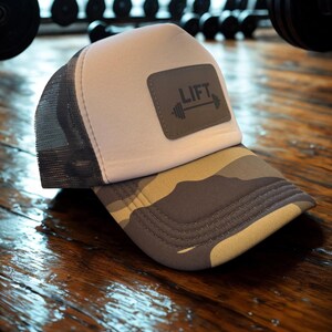May include: A grey and white trucker hat with a camouflage brim. The hat features a rectangular patch with the word "LIFT" and a barbell graphic. The hat is designed for fitness enthusiasts.