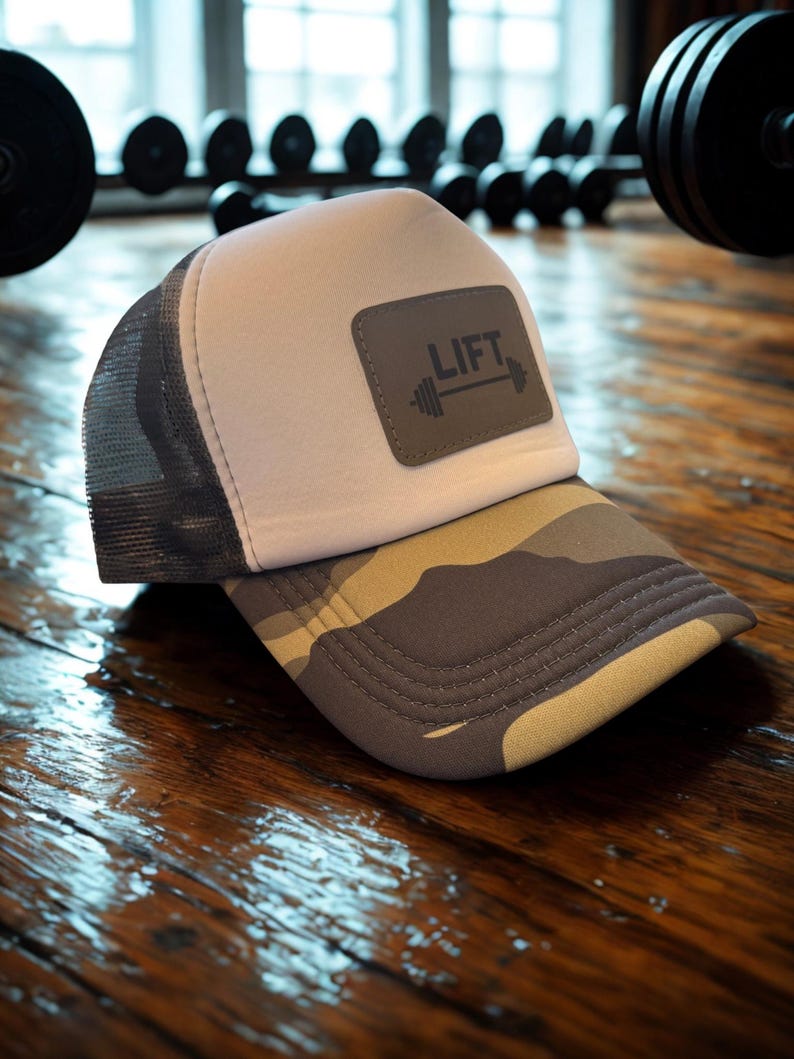 May include: A grey and white trucker hat with a camouflage brim. The hat features a rectangular patch with the word "LIFT" and a barbell graphic. The hat is designed for fitness enthusiasts.