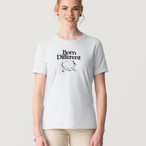 May include: A light gray short-sleeved t-shirt featuring the black text Born Different printed on the chest above a black line drawing of a small, stout creature walking on all fours. The shirt has a crew neckline and a standard fit.