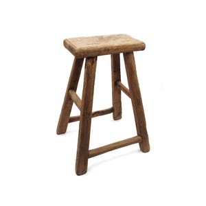 Könnte beinhalten: Ein rustikaler Holzschemel mit rechteckiger Sitzfläche und vier abgewinkelten Beinen. Der Schemel besteht aus verwittertem Holz und zeigt natürliche Maserung und Knoten. Das Design ist einfach und funktional, mit Fokus auf die natürliche Schönheit des Materials.