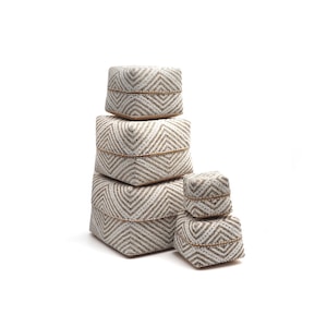 Puede incluir: Conjunto de cuatro cajas de almacenamiento cuadradas de material tejido, de color blanco y marrón, con un patrón geométrico. Las cajas están apiladas unas sobre otras.