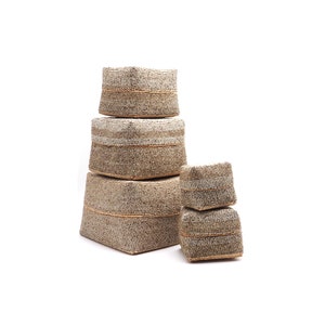 May include: Set of four square storage boxes made of woven beads. The boxes are stacked in a pyramid shape, with the largest box at the bottom and the smallest box at the top. The boxes are a light brown color with a subtle shimmer.