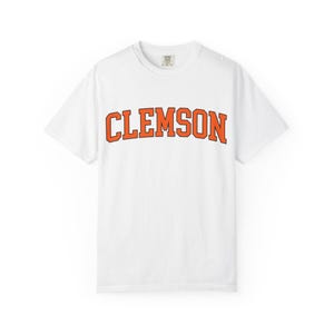 May include: White t-shirt with the word "CLEMSON" printed in orange block letters across the chest. The shirt has a crew neck and short sleeves. The fabric appears to be a soft cotton blend.