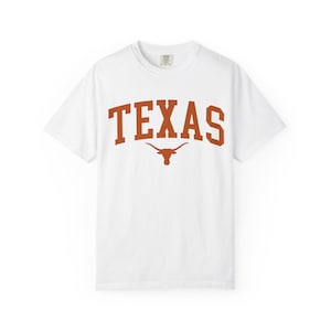 May include: White t-shirt with the word "TEXAS" in burnt orange block letters and a longhorn logo. The shirt has a crew neck and short sleeves, perfect for casual wear.
