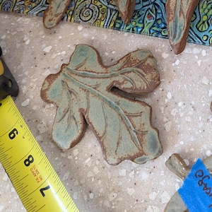 Puede incluir: Una colección de adornos de cerámica con forma de hoja que presentan un esmalte verde claro con detalles marrones. Las hojas varían en forma y tamaño, con una cinta métrica amarilla visible en la esquina que muestra medidas en centímetros.