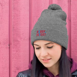May include: A grey beanie with a pom pom on top. The beanie has the words "Merry Bright" embroidered on the front in red and blue.