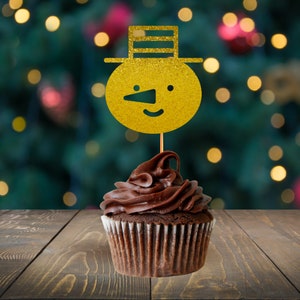 May include: A chocolate cupcake with chocolate frosting, decorated with a gold glitter snowman cake topper. The snowman has a black nose and eyes, and a black hat. The cupcake is on a wooden surface with a blurred Christmas lights background.