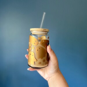 Op de afbeelding: Een heldere glazen drankbeker met een bamboe deksel en rietje, gevuld met een lichtbruine vloeistof. Het glas is versierd met gouden spookstickers. De achtergrond is effen blauw.
