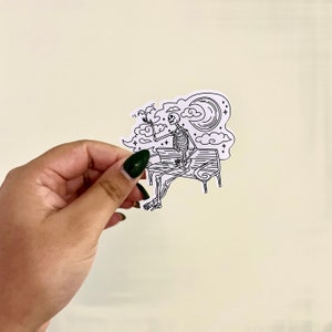 Puede incluir: Un dibujo en blanco y negro de un esqueleto sentado en un banco con una luna creciente y nubes en el fondo.