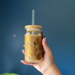 Op de afbeelding: Een heldere glazen beker met een bamboe deksel en een doorzichtig rietje. De beker is versierd met gouden spook- en sterontwerpen. Het glas is gevuld met een lichtbruine vloeistof.