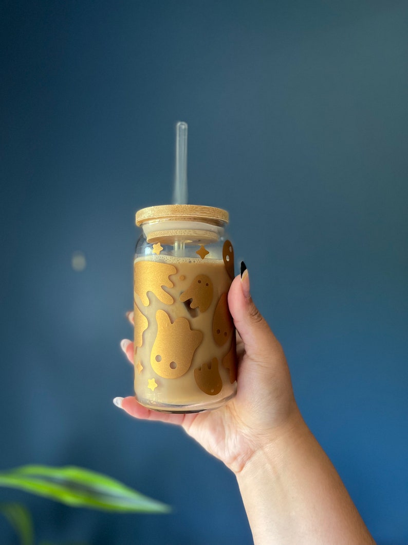 Op de afbeelding: Een heldere glazen beker met een bamboe deksel en een doorzichtig rietje. De beker is versierd met gouden spook- en sterontwerpen. Het glas is gevuld met een lichtbruine vloeistof.