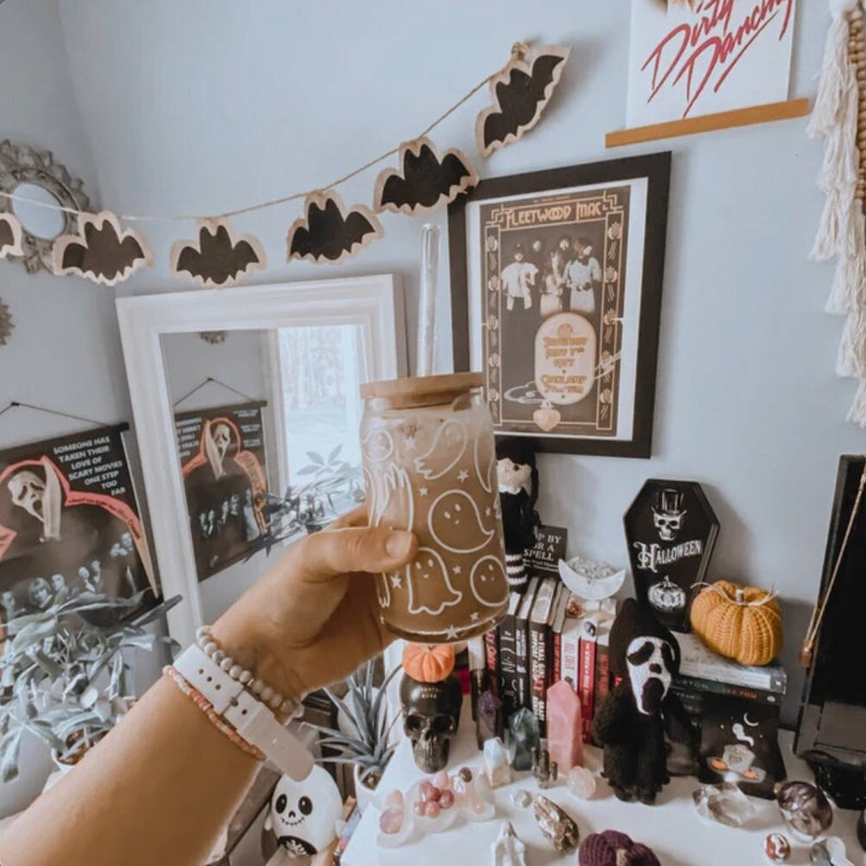 Puede incluir: Un tarro de vidrio blanco con una tapa marr&oacute;n y un dise&ntilde;o de fantasma. El tarro est&aacute; lleno de un l&iacute;quido marr&oacute;n claro. El tarro est&aacute; sentado en una mesa con una variedad de decoraciones de Halloween, incluyendo una calavera, una calabaza y una figura de fantasma. Las decoraciones est&aacute;n en una mesa con una superficie blanca. Hay una cadena de murci&eacute;lagos de papel negro colgando del techo.