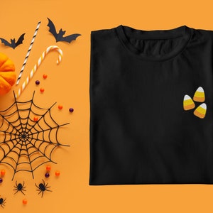 Peut inclure: T-shirt noir avec trois applications de maïs sucré orange et jaune sur le devant. Le t-shirt est sur un fond orange avec une toile d'araignée, du maïs sucré, des citrouilles et des chauves-souris.