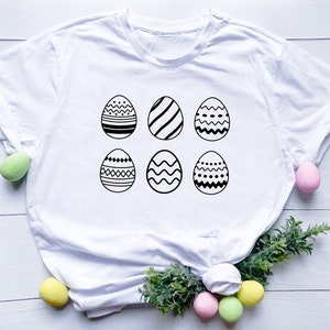 Peut inclure: T-shirt blanc avec six motifs d'œufs de Pâques noirs et blancs. Les œufs sont disposés en deux rangées de trois.