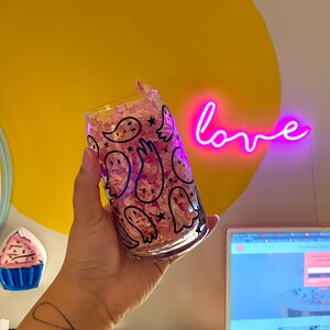 Puede incluir: Un vaso transparente con un patr&oacute;n de fantasmas en blanco y negro. El vaso est&aacute; lleno de papel de seda rosa y est&aacute; siendo sostenido por una mano. El fondo es amarillo con un letrero de ne&oacute;n rosa que dice "love".