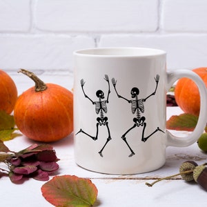 May include: White ceramic coffee mug featuring a black skeleton design. The skeletons are dancing with raised arms. The mug is on a white wooden surface with autumn leaves and pumpkins.