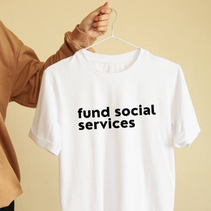 May include: White t-shirt with black text that reads "fund social services".
