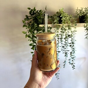 Op de afbeelding: Een heldere glazen beker gevuld met een lichtbruine drank, met een bamboe deksel en een helder glazen rietje. Het glas is versierd met goudkleurige spookontwerpen. De beker wordt vastgehouden voor een achtergrond van groene planten.