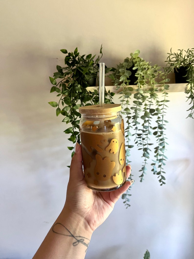 Op de afbeelding: Een heldere glazen beker gevuld met een lichtbruine drank, met een bamboe deksel en een helder glazen rietje. Het glas is versierd met goudkleurige spookontwerpen. De beker wordt vastgehouden voor een achtergrond van groene planten.