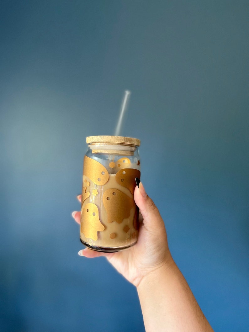 Op de afbeelding: Een heldere glazen drankbeker met een bamboe deksel en rietje, gevuld met een lichtbruine vloeistof. Het glas is versierd met gouden spookstickers. De achtergrond is effen blauw.