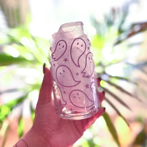 Puede incluir: Un vaso de vidrio transparente con un dise&ntilde;o rosa transl&uacute;cido que presenta un patr&oacute;n repetitivo de fantasmas blancos y estrellas. El vaso est&aacute; siendo sostenido por una mano.