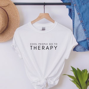 May include: White t-shirt with black text that reads "Cool People Go To Therapy".