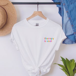 May include: White t-shirt with the text "therapy is cool" in colourful letters on the front.
