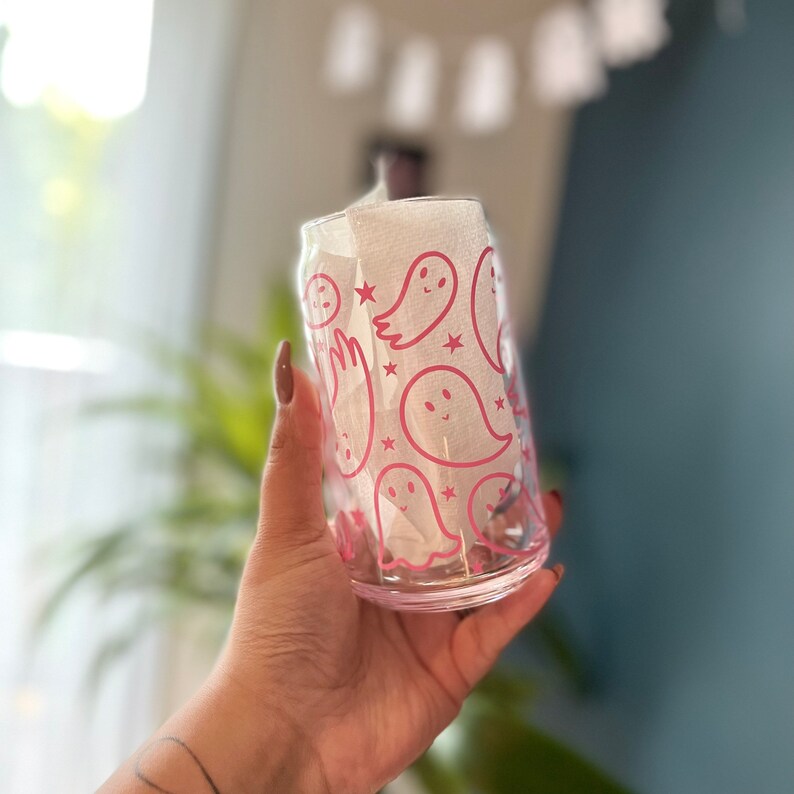 Puede incluir: Un vaso de vidrio transparente con un dise&ntilde;o de fantasmas rosas. El vaso tiene un contorno rosa de fantasmas con estrellas y una servilleta de papel blanca en su interior.