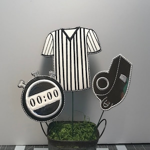 Könnte beinhalten: Ein schwarz-weißes Schiedsrichtertrikot, eine Pfeife und eine Stoppuhr mit der Anzeige 00:00, alles in einem Metallkübel mit grünem Moos angeordnet.