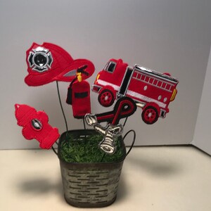 May include: A decorative arrangement featuring felt cutouts of a red fire truck, fire hat, fire hydrant, fire extinguisher, and a fire axe. The cutouts are arranged in a small, galvanized metal bucket filled with green moss.