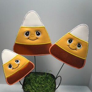 May include: Three felt candy corn decorations in a small metal bucket with green moss. The candy corn are orange, yellow, and white with embroidered eyes and a smiling mouth.