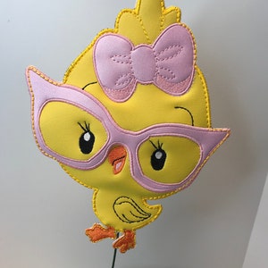 May include: A yellow chick with pink glasses and a pink bow on its head. The chick is standing on a green stem.
