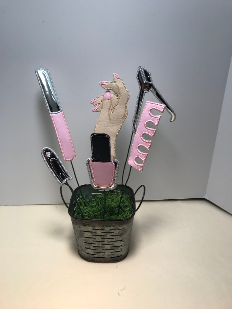 K&ouml;nnte beinhalten: Ein Metalleimer gef&uuml;llt mit gr&uuml;nem Moos, obenauf eine Hand und verschiedene Nagelpflegewerkzeuge, darunter eine Nagelfeile, Nagelknipser und Nagellack.