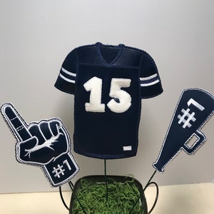 May include: A football-themed centerpiece with a blue jersey with the number 15, a blue foam finger with the number 1, and a blue megaphone with the number 1. The items are arranged in a small metal bucket with green moss.