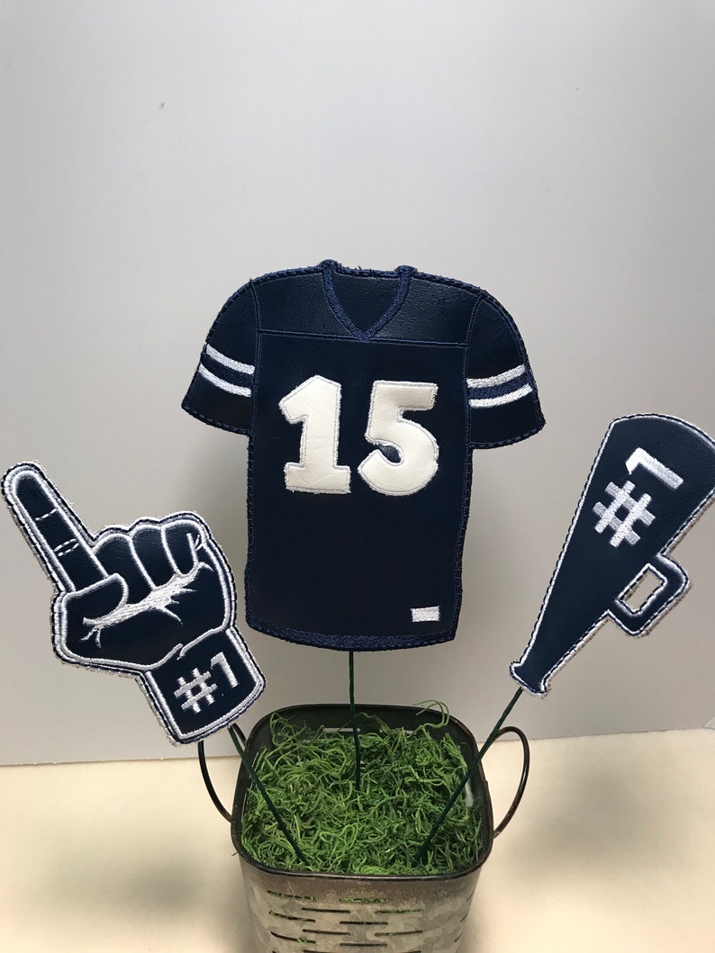 May include: A football-themed centerpiece with a blue jersey with the number 15, a blue foam finger with the number 1, and a blue megaphone with the number 1. The items are arranged in a small metal bucket with green moss.